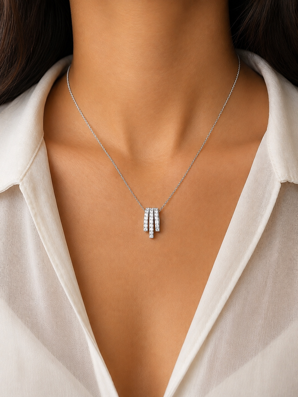 A close-up of a model wearing a delicate silver necklace with a triple-bar pendant set with small clear stones, paired with a white collared blouse.