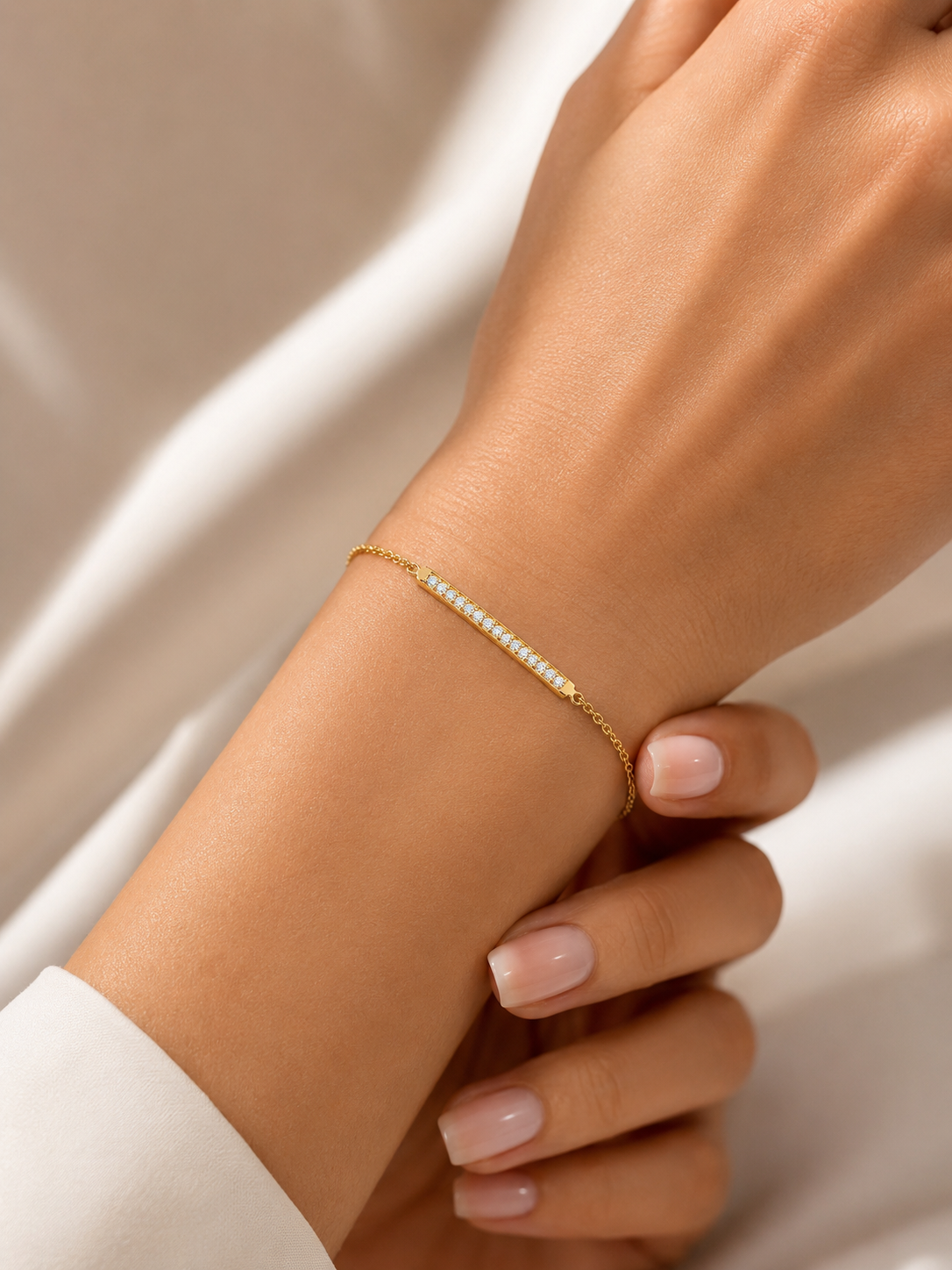 A delicate gold chain bracelet with a row of small lab diamond, worn on a person's wrist against a soft white fabric background.